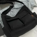 Trendy black oversized messenger bag placed beside laptop and denim outfit