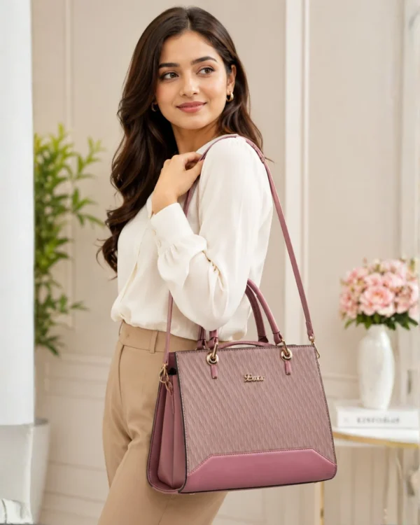 Elegant pink structured handbag for women carried by a model, featuring dual top handles, detachable shoulder strap, textured finish, and gold-tone hardware.