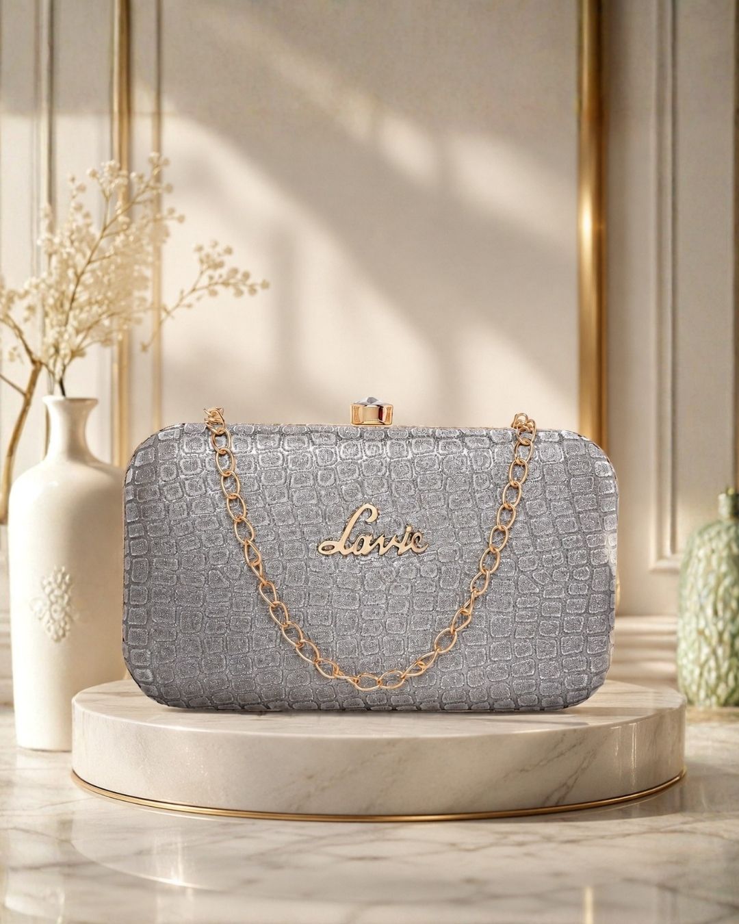 Luxury silver textured clutch bag with a gold chain strap displayed on a premium beige pedestal in an elegant studio setting.